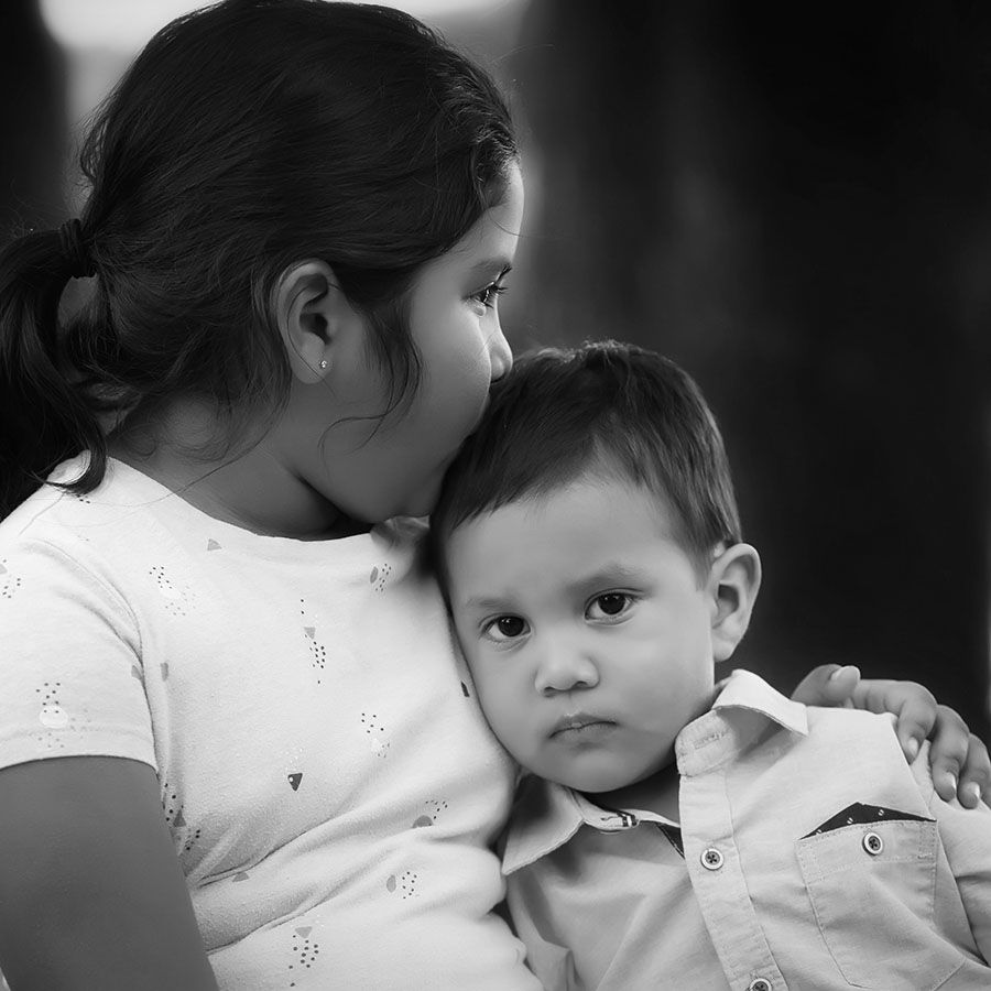 pensive young hispanic children. older girl has her arm around younger boy while she kisses his forehead