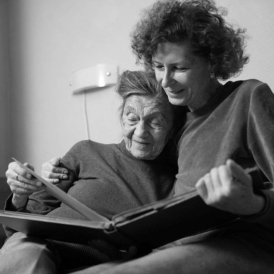 middle-aged woman sitting on a couch with her arm around an older woman while they look at a photo album